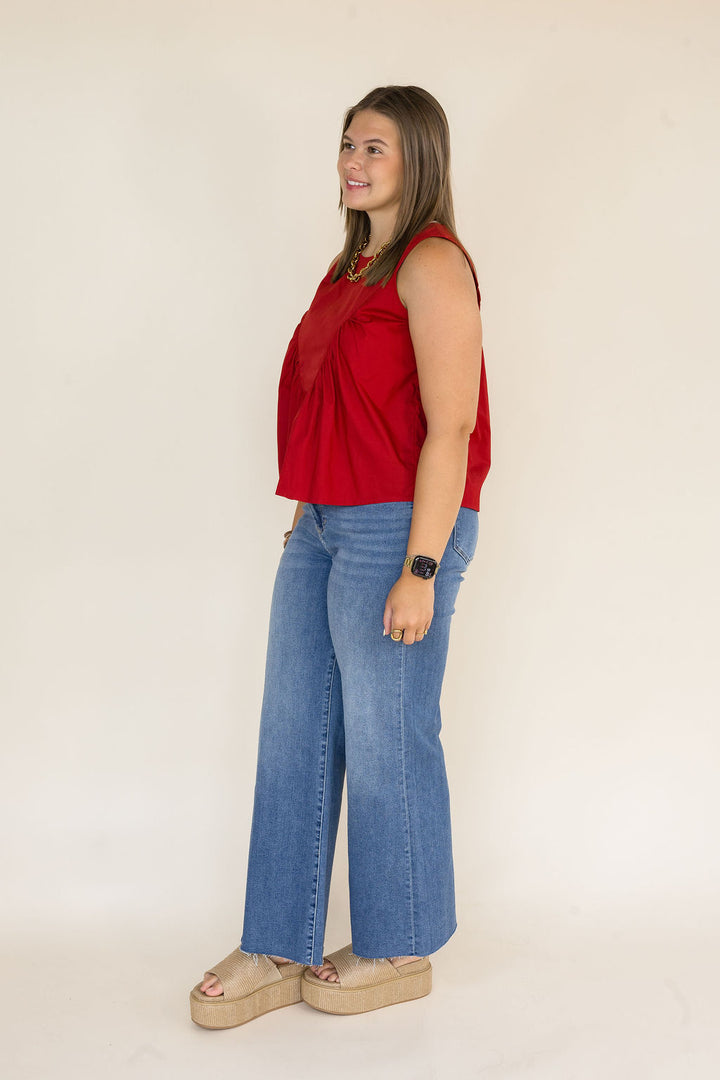A woman modeling a sleeveless garnet top and wide-leg blue jeans with beige platform sandals, standing against a plain light beige studio background.