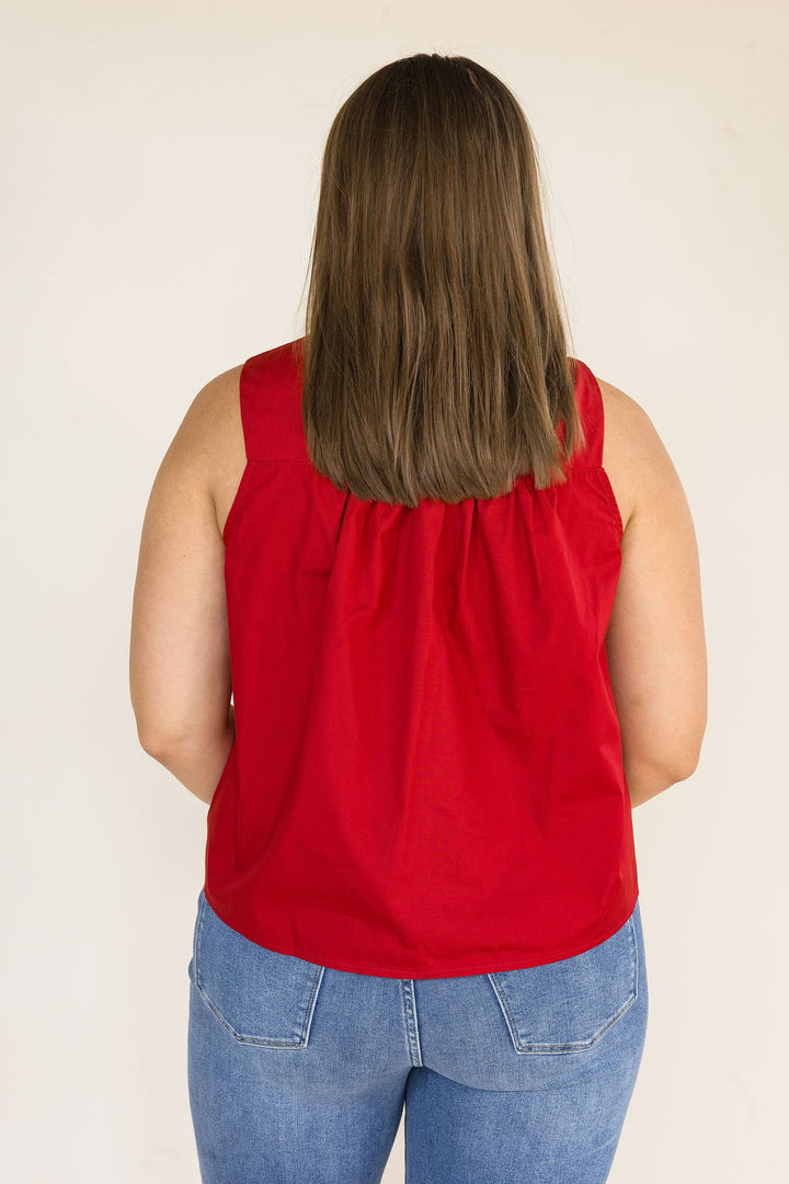 A woman viewed from behind wearing a garnet sleeveless top and blue jeans, standing against a plain off-white background.