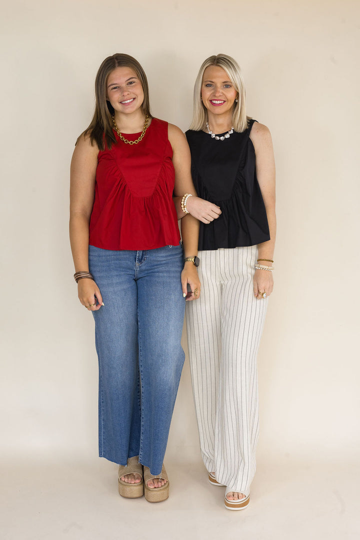 Two women standing side-by-side modeling sleeveless peplum tops. The woman on the left wears a garnet top with wide-leg blue jeans, and the woman on the right wears a black top with cream pinstriped pants.