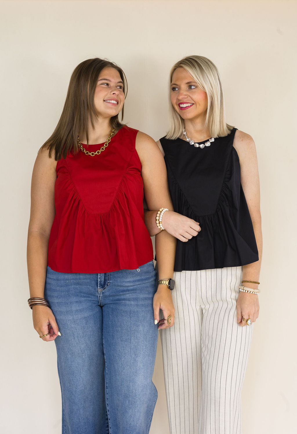 Two women standing side-by-side wearing sleeveless peplum tops, one in garnet and one in black, paired with jeans and pinstriped pants against a plain beige background.