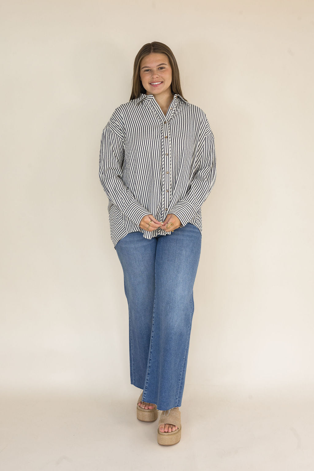 Woman wearing a navy and ivory striped oversized button-down shirt featuring a collared neckline, tortoise buttons, long sleeves, and a relaxed high-low hem paired with light wash wide leg jeans.