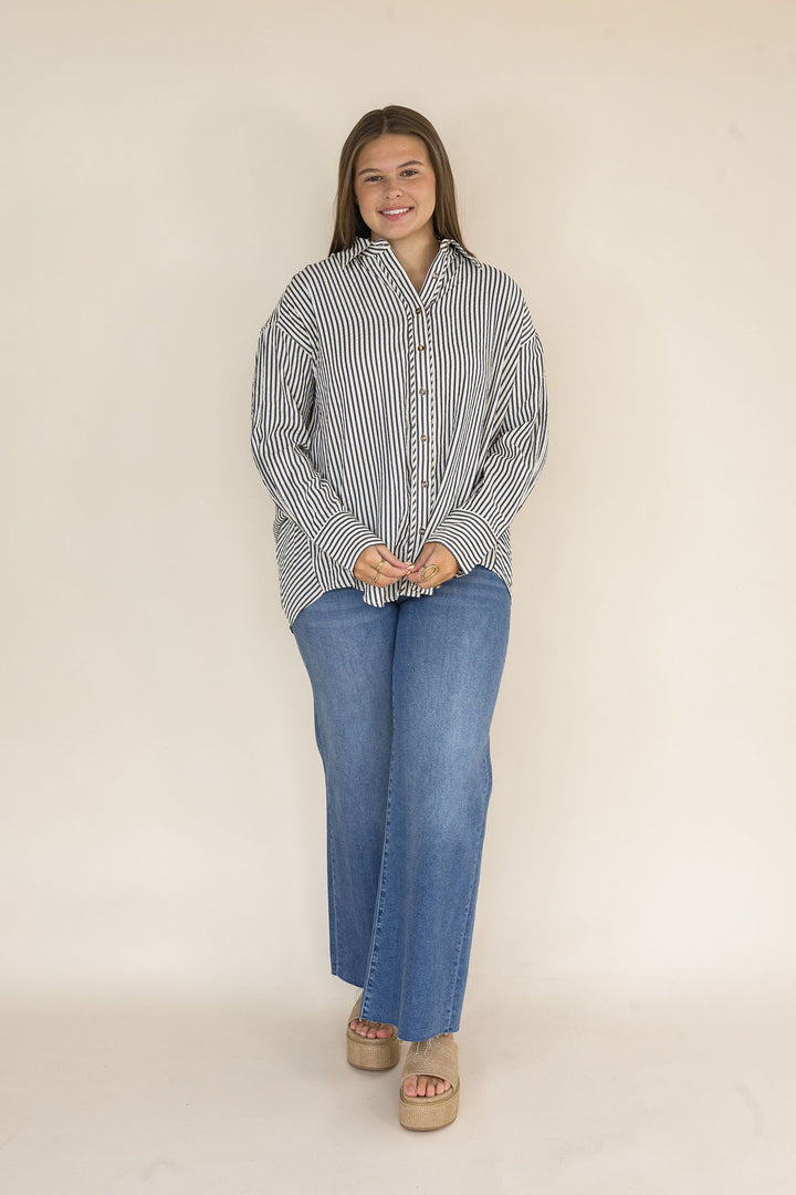 Woman wearing a navy and ivory striped oversized button-down shirt featuring a collared neckline, tortoise buttons, long sleeves, and a relaxed high-low hem paired with light wash wide leg jeans.