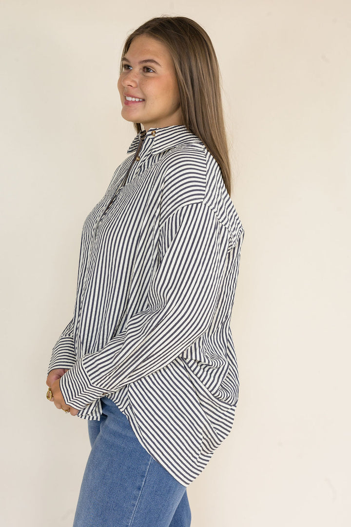 Side view of a woman wearing a navy and ivory striped oversized button-down shirt with a collared neckline, long sleeves, and a curved high-low hem paired with light wash wide leg jeans.