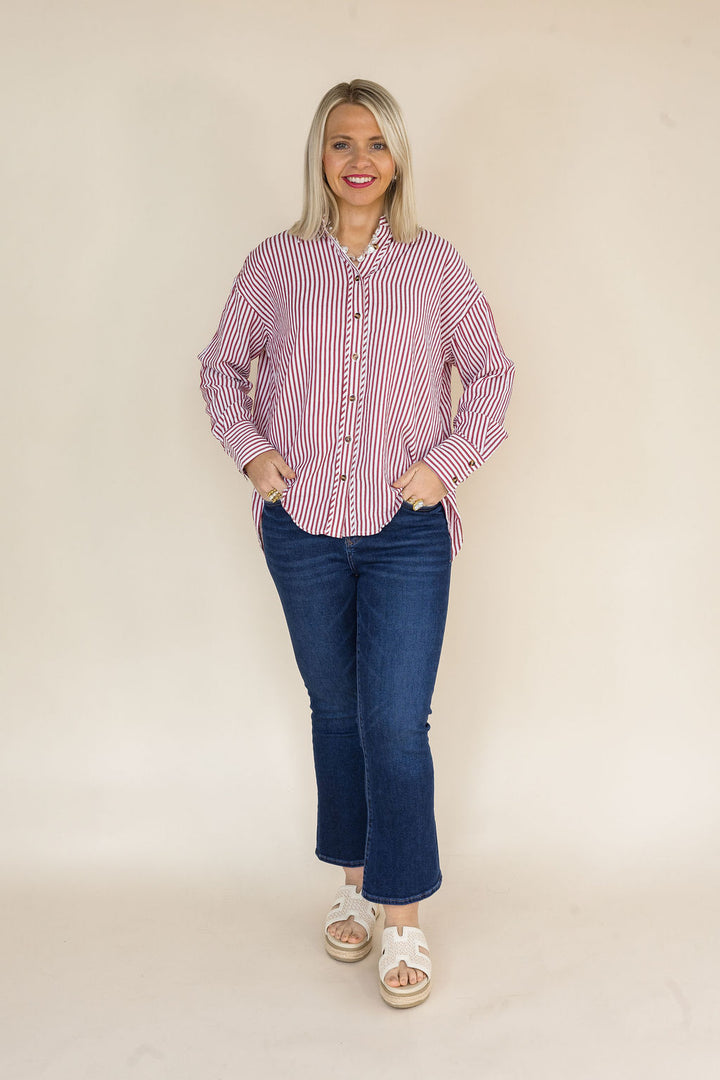 A woman modeling a brick and white vertically striped button-down shirt with dark blue jeans and white slide sandals, standing against a plain beige studio backdrop.