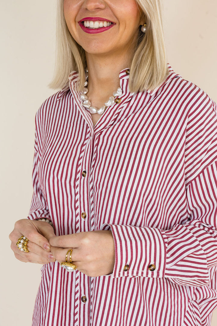 A woman wearing a brick and white vertically striped button-down shirt, styled with a pearl necklace and gold rings, against a plain beige background.
