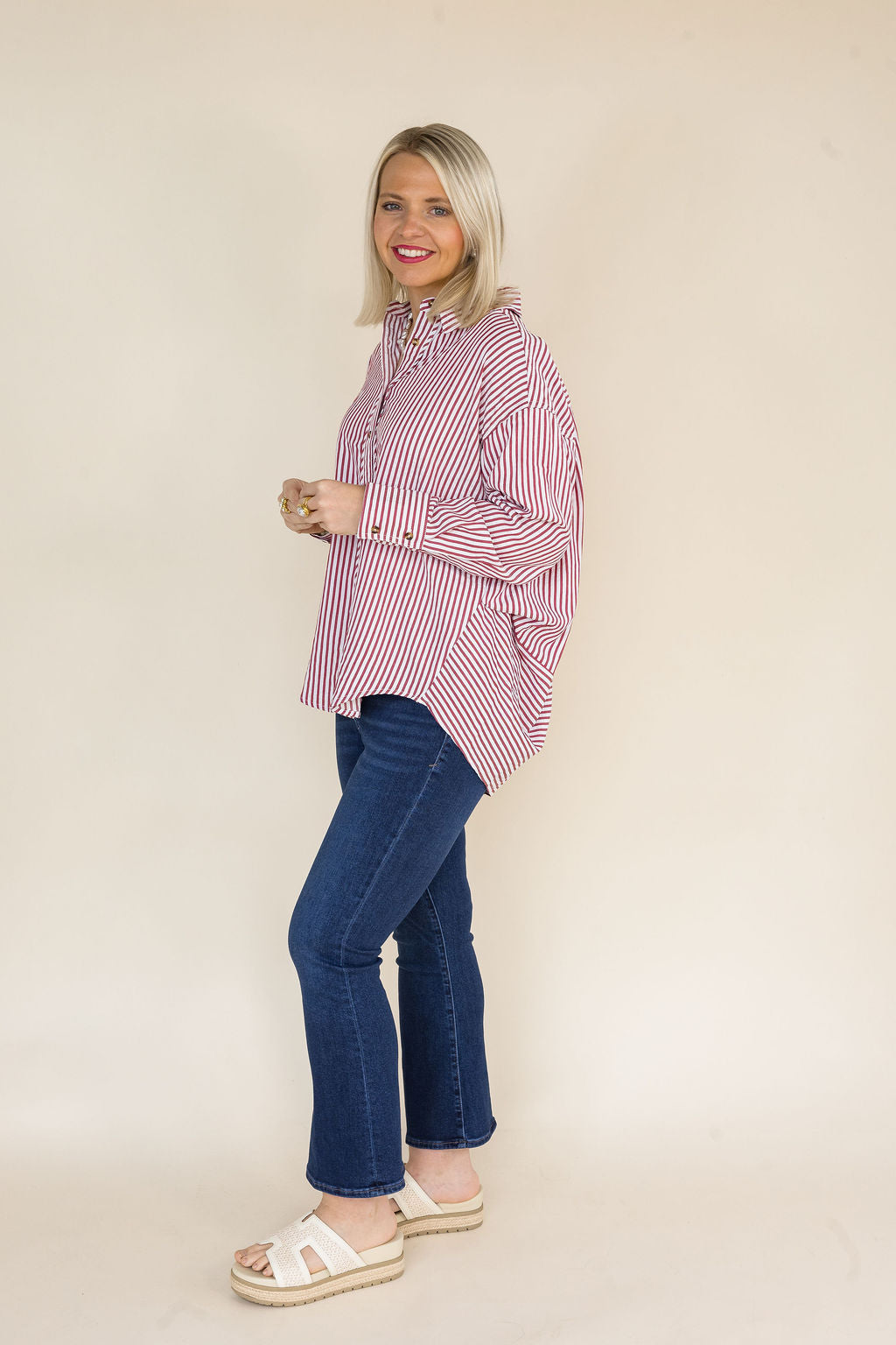 Side view of a woman wearing a red and white striped oversized button-down shirt featuring a collared neckline, long sleeves with button cuffs, and a relaxed high-low hem paired with dark wash cropped flare jeans and ivory platform slide sandals.