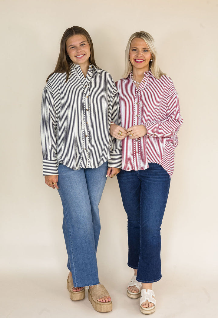 Two women modeling long-sleeve button-up shirts with vertical stripes, standing against a plain beige studio background. The woman on the left wears a dark navy and white striped shirt with blue jeans, and the woman on the right wears a brick and white striped shirt with blue jeans.