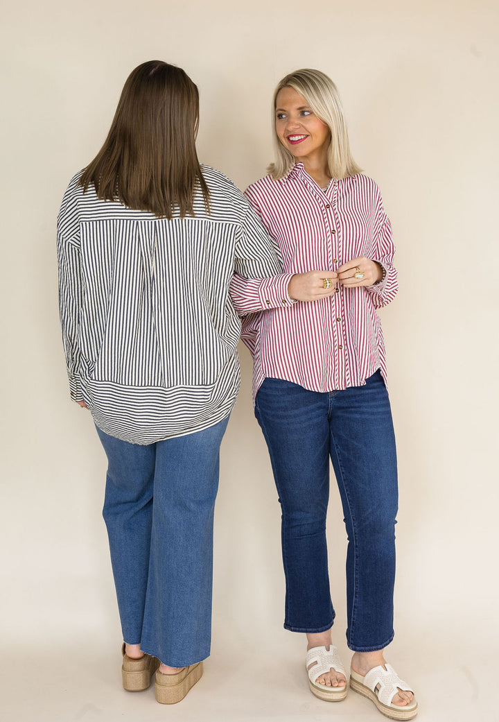 Two women modeling striped button-up shirts and jeans against a plain beige studio background. The woman on the left wears a navy-and-white vertically striped shirt with blue jeans, and the woman on the right wears a brick-and-white vertically striped shirt with blue jeans.