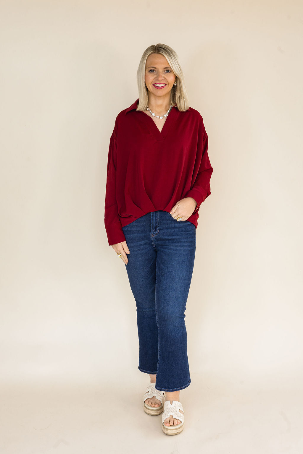 Woman wearing a burgundy oversized V-neck collared blouse with long sleeves, drop shoulders, and dark wash cropped flare jeans styled with ivory platform slide sandals.