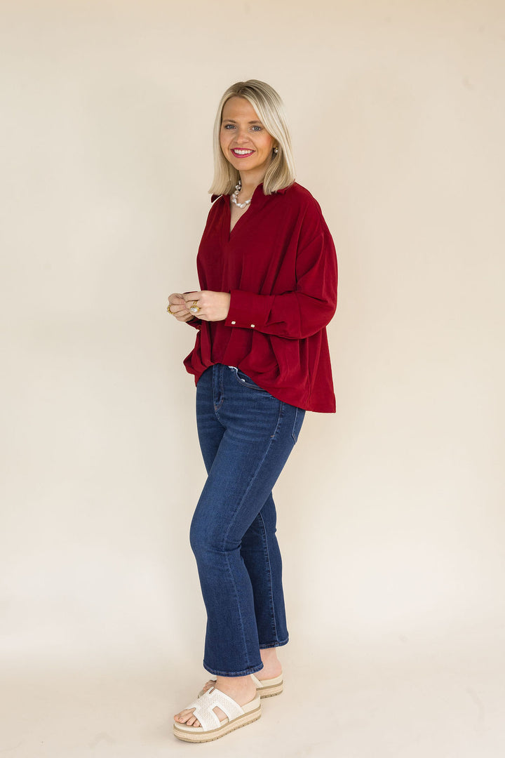 Side view of a woman wearing a burgundy oversized collared blouse with a V-neckline, long sleeves, and a relaxed silhouette paired with dark wash cropped flare jeans.