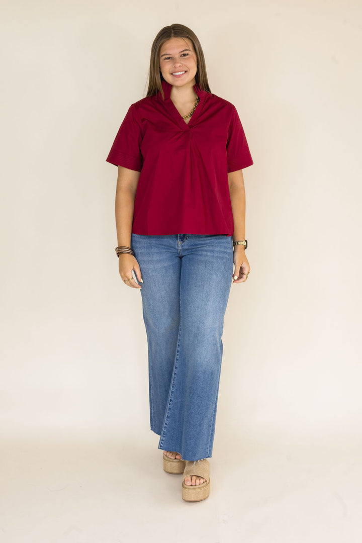 A woman modeling a short-sleeve, gathered-neck blouse in deep burgundy, paired with wide-leg blue jeans and beige platform sandals, standing against a plain beige studio backdrop.