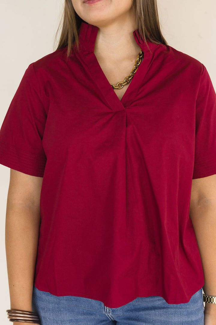 A woman wearing a short-sleeve top with a gathering detail at the collar, in a deep burgundy color, paired with blue jeans and a gold chain necklace