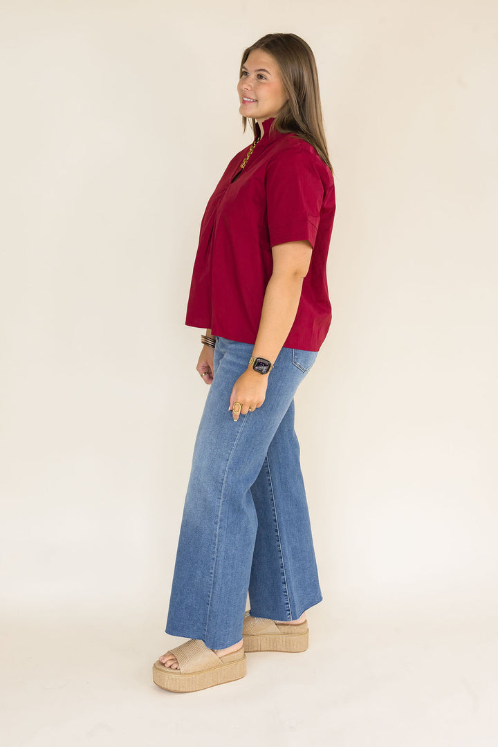 A woman modeling a short-sleeve burgundy top and wide-leg blue jeans with beige platform sandals, shown in profile against a plain off-white studio background.