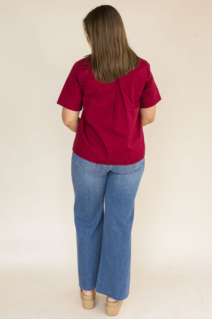 A woman viewed from behind wearing a short-sleeve burgundy blouse and blue jeans, standing against a plain off-white studio background.