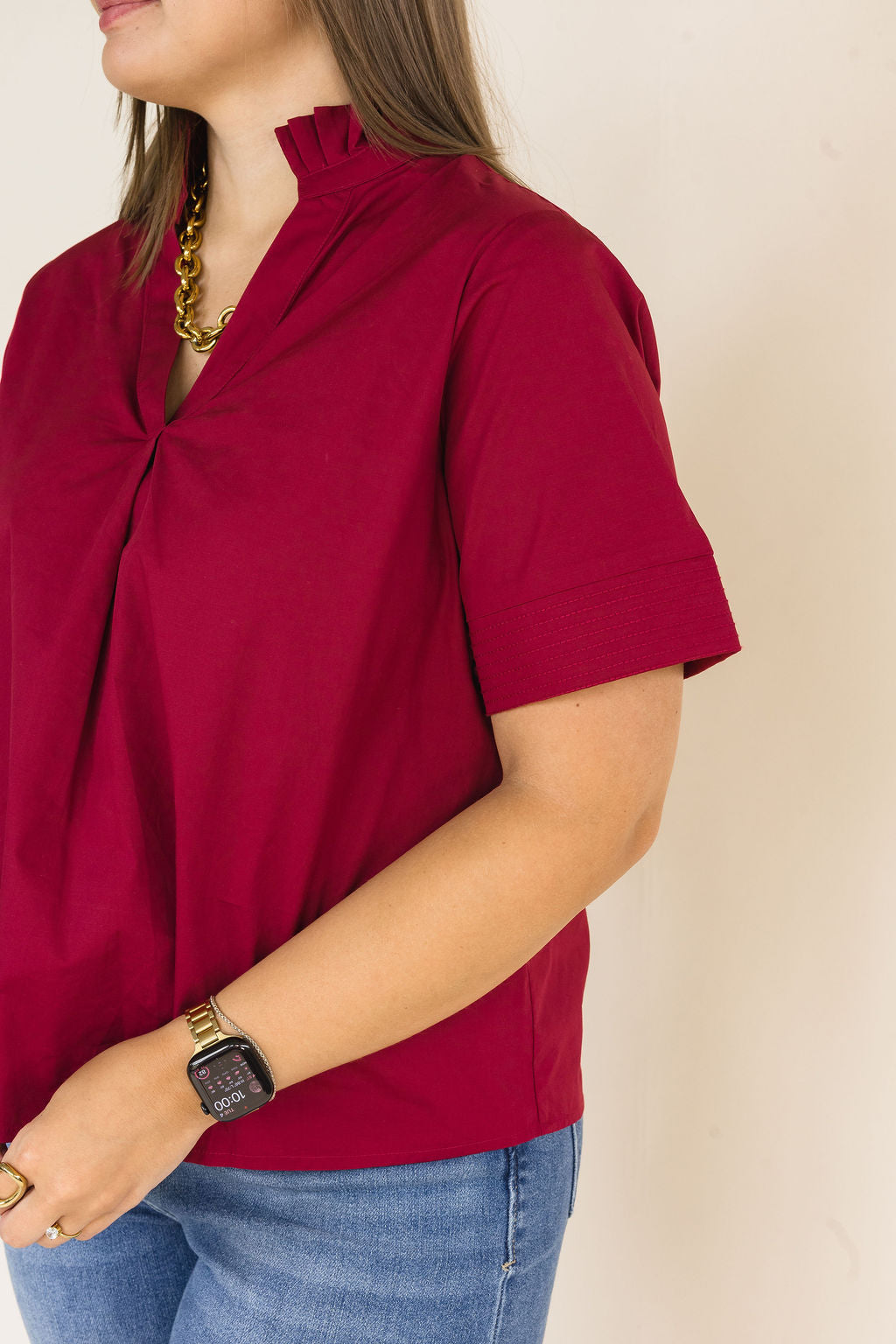 A woman wearing a short-sleeve, high-neck blouse in deep burgundy with a gathered front detail, paired with blue jeans and a gold chain necklace, shown against a plain beige background.