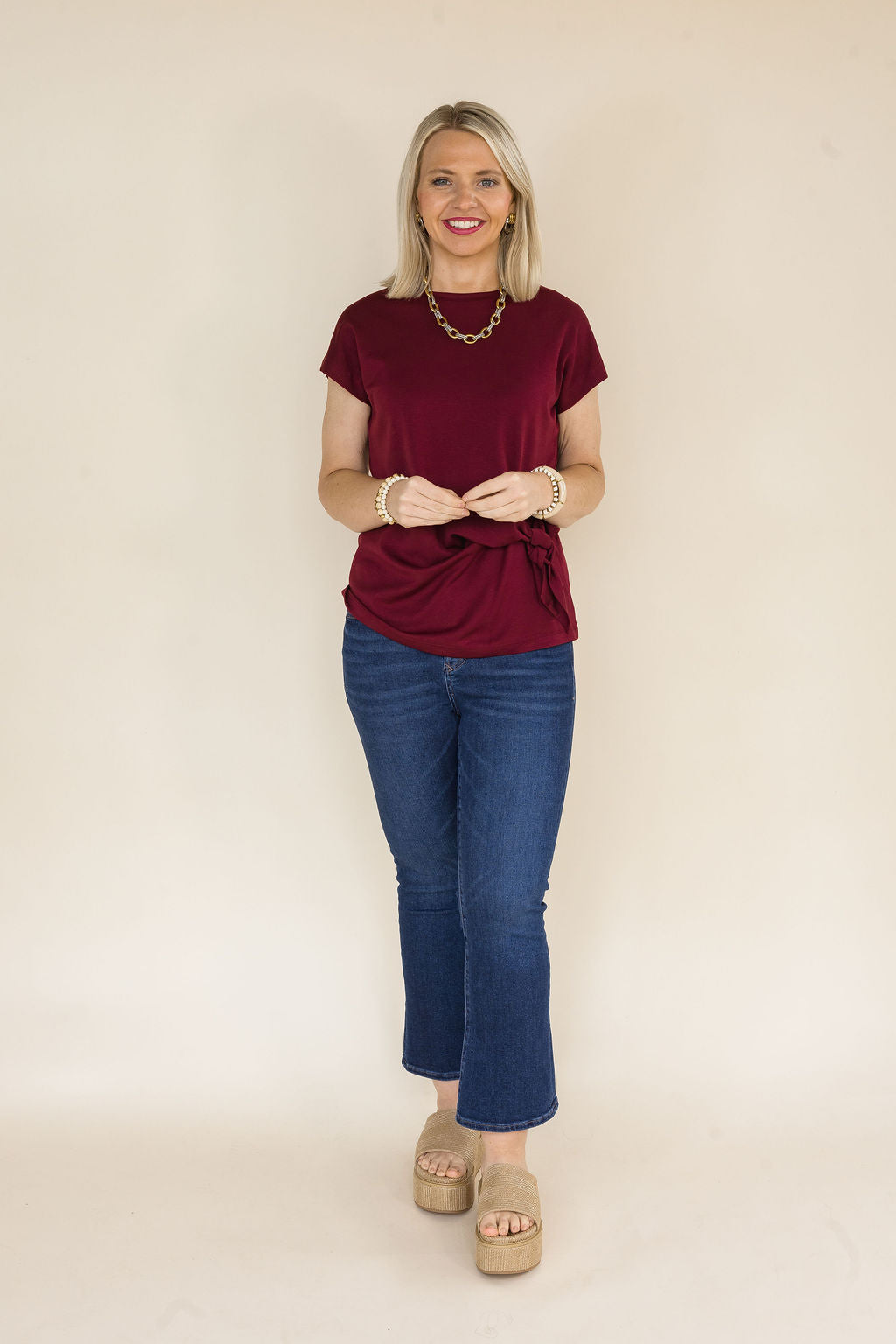 A woman modeling a short-sleeve maroon blouse with a tie waist detail, paired with blue jeans and beige platform sandals, standing against a plain beige background.