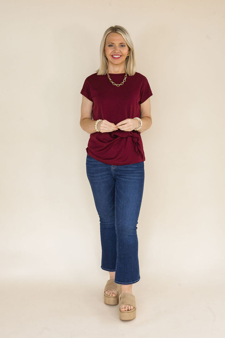 A woman modeling a short-sleeve maroon blouse with a tie waist detail, paired with blue jeans and beige platform sandals, standing against a plain beige background.