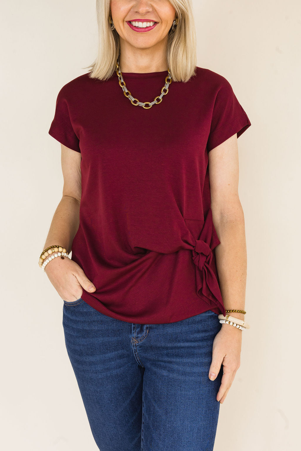 A woman wearing a short-sleeve maroon blouse with a side tie detail, paired with blue jeans, against a plain light beige background.