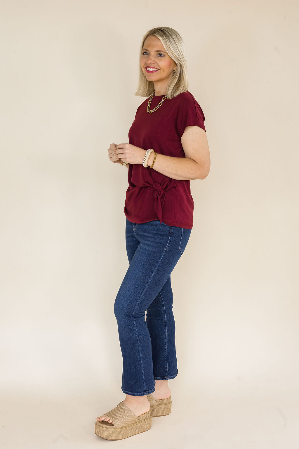 A woman modeling a short-sleeve maroon blouse with a side tie detail, paired with blue jeans and beige platform sandals, standing against a plain beige studio background.