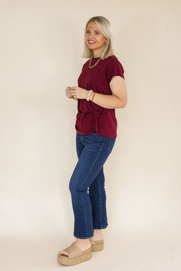 A woman modeling a short-sleeve maroon blouse with a side tie detail, paired with blue jeans and beige platform sandals, standing against a plain beige studio background.