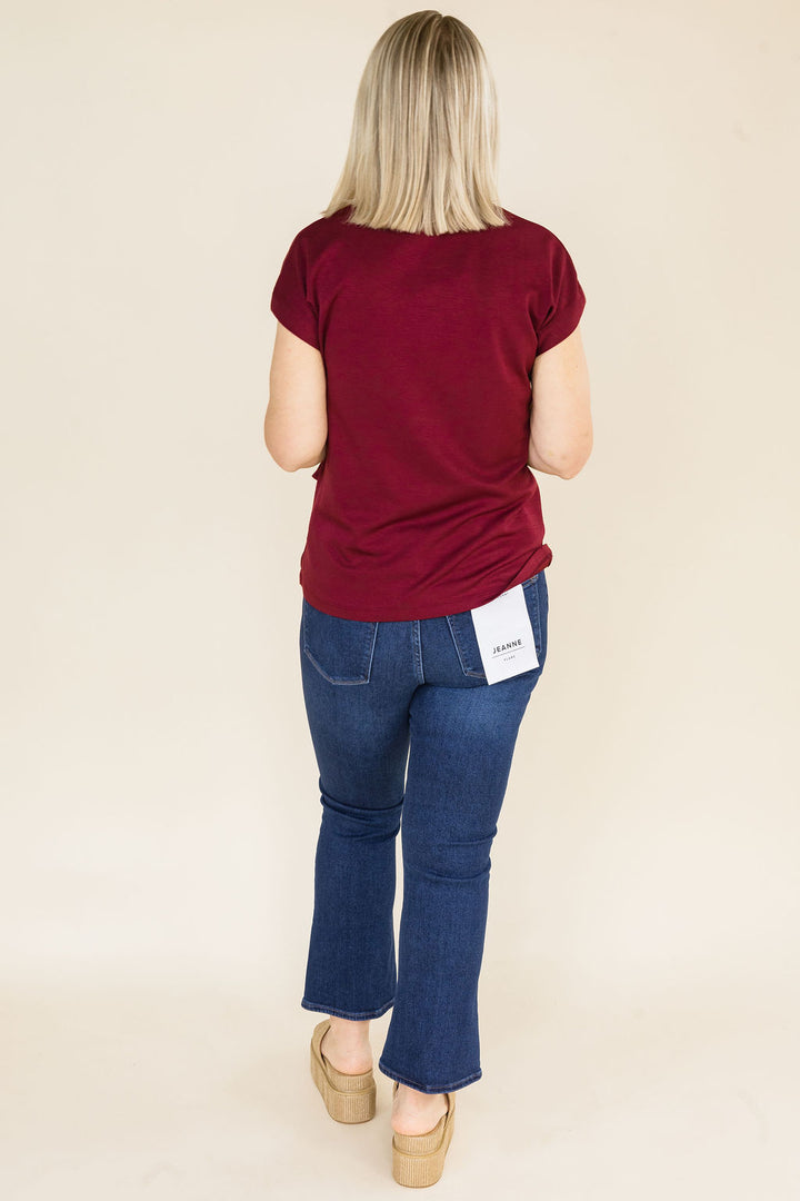 A woman viewed from behind wearing a short-sleeve burgundy blouse and blue jeans, standing against a plain beige studio background.