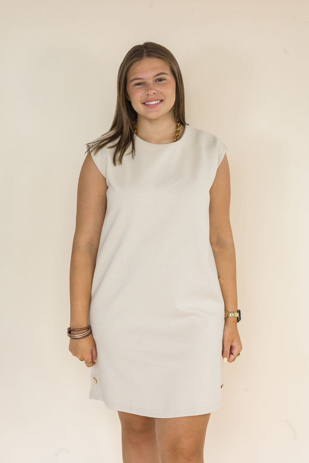 A woman modeling a sleeveless, knee-length off-white dress against a plain, warm beige background.