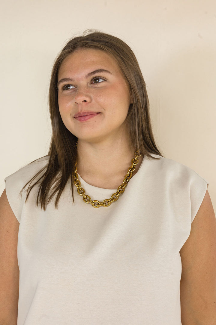 A young woman with brown hair wearing a chunky gold chain necklace and a cream-colored sleeveless dress, looking off to the side against a plain beige wall.