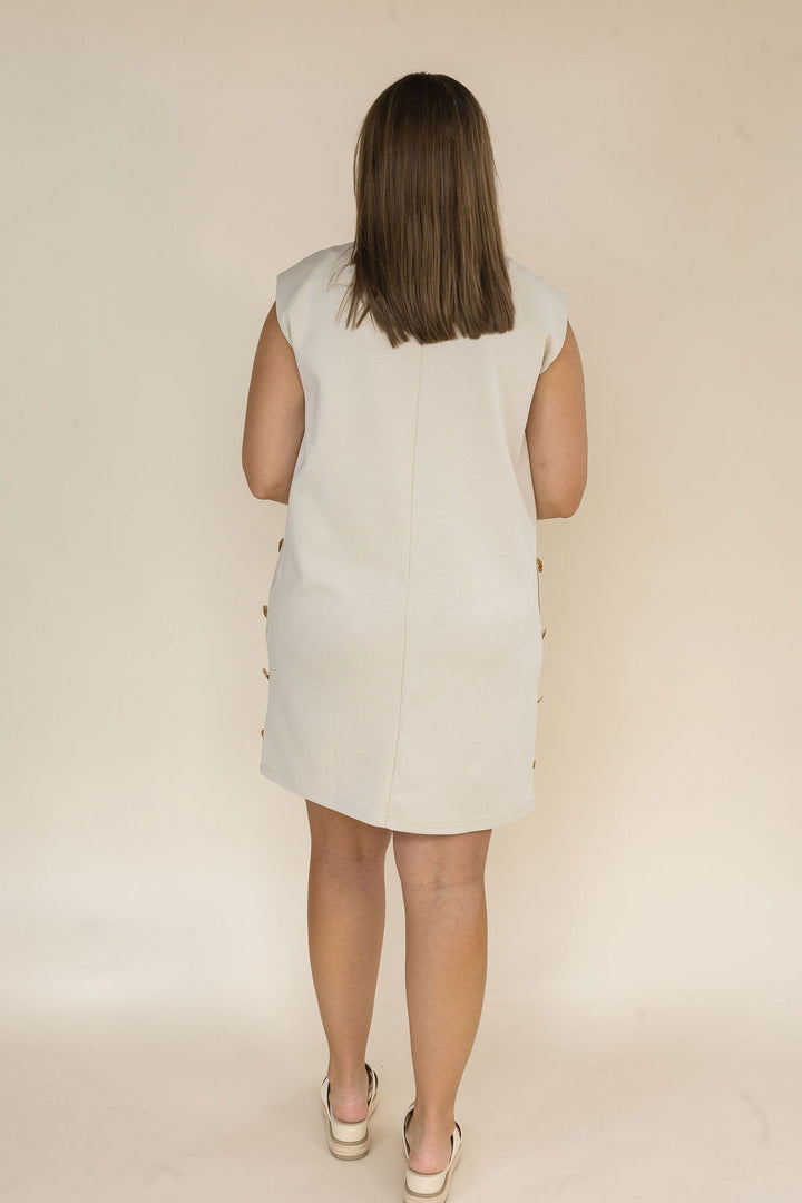 A woman viewed from behind wearing a sleeveless cream-colored dress with gold buttons on the sides, standing against a plain beige studio background.