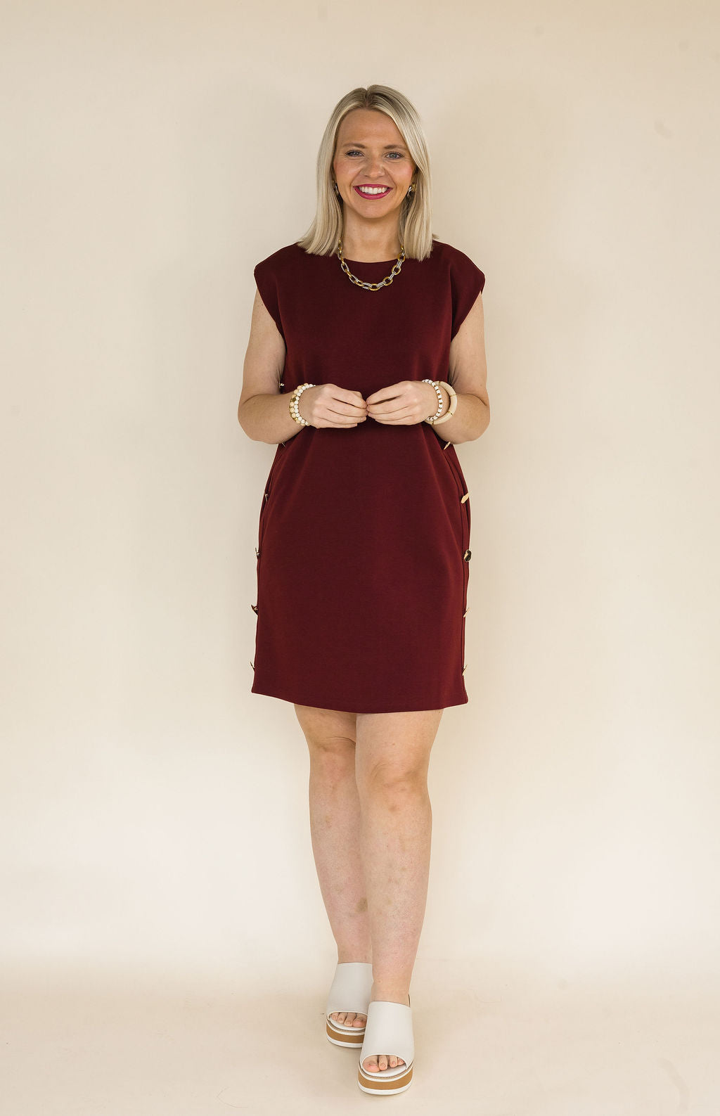 Woman wearing a burgundy sleeveless shift dress featuring decorative gold side buttons, side pockets, and a classic crew neckline styled with ivory platform mule heels.