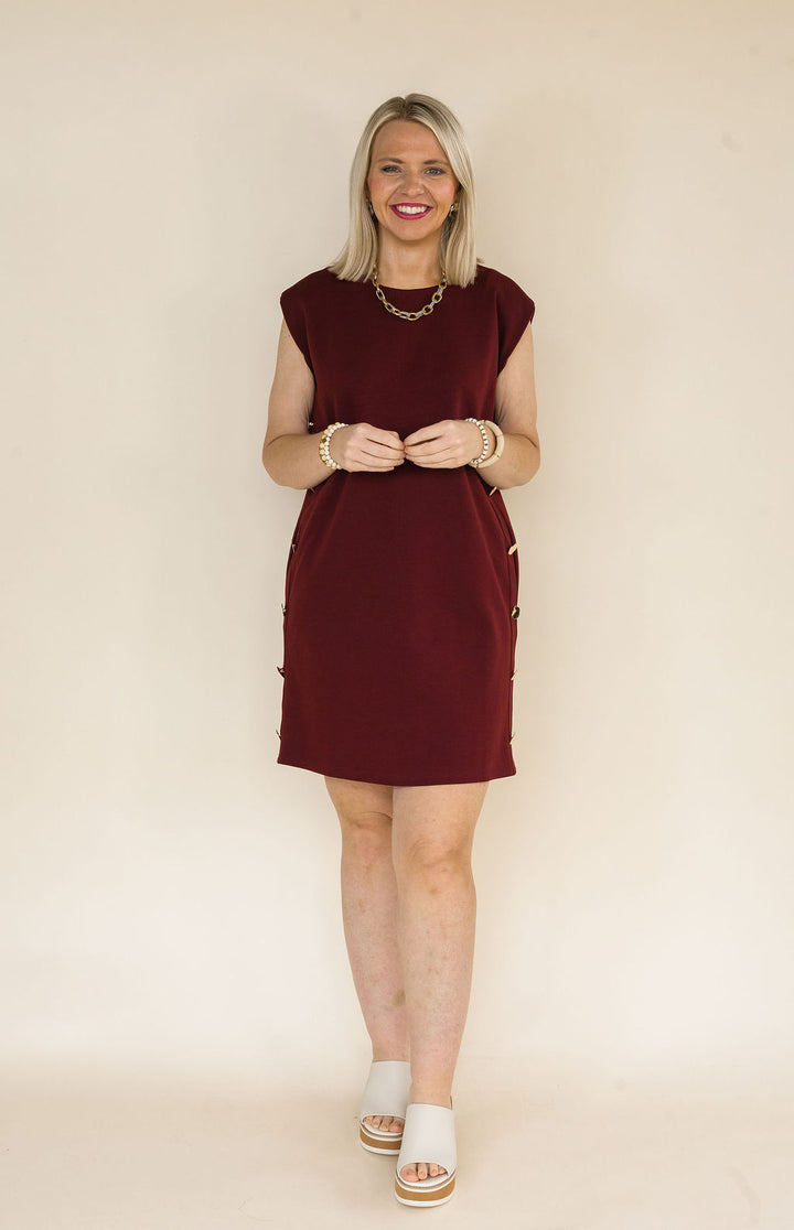 Woman wearing a burgundy sleeveless shift dress featuring decorative gold side buttons, side pockets, and a classic crew neckline styled with ivory platform mule heels.