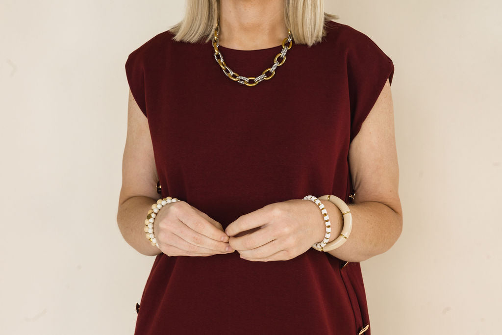 A woman wearing a sleeveless burgundy dress and gold chain necklace, accessorized with pearl and beige beaded bracelets, against a plain beige background.