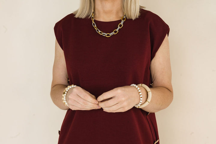 A woman wearing a sleeveless burgundy dress and gold chain necklace, accessorized with pearl and beige beaded bracelets, against a plain beige background.