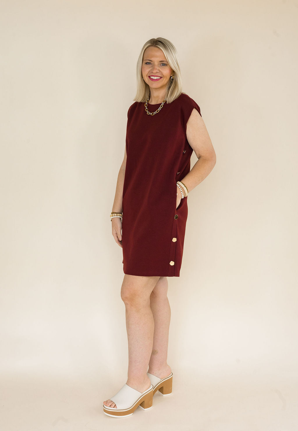 A woman modeling a sleeveless burgundy shift dress with gold buttons down the side, standing against a plain beige studio background.