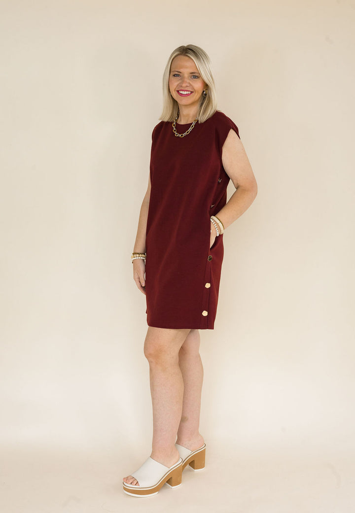 A woman modeling a sleeveless burgundy shift dress with gold buttons down the side, standing against a plain beige studio background.