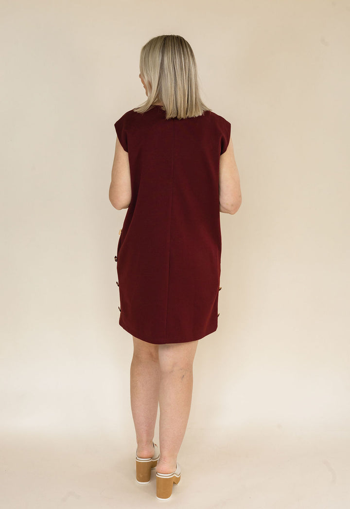 A woman with blonde hair is shown from behind wearing a sleeveless burgundy dress with decorative buttons down the sides, standing against a plain beige background.