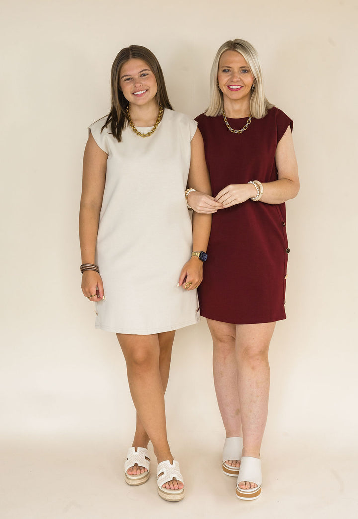 Two women standing side-by-side wearing sleeveless mini dresses, one in cream and the other in burgundy, against a plain cream studio background.