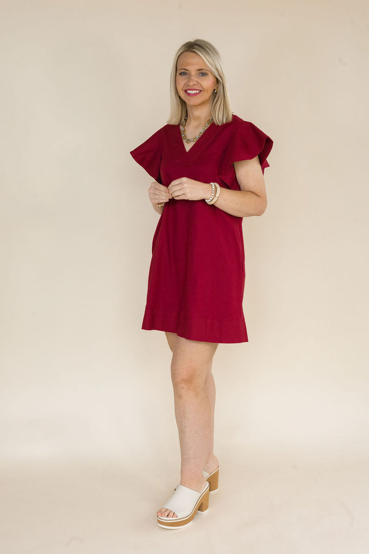 Side view of a woman wearing a burgundy flutter sleeve shift dress with a V-neckline, textured fabric, and an easy relaxed fit paired with ivory platform mule heels.