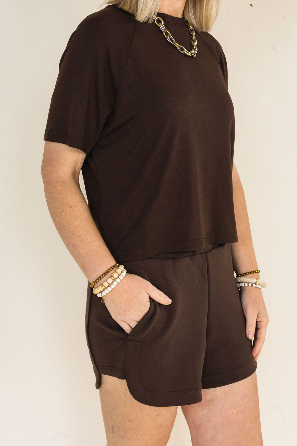 A woman wearing a coffee short-sleeve t-shirt and matching coffee shorts with side pockets, accessorized with gold chain and pearl bracelets, against a plain off-white background.