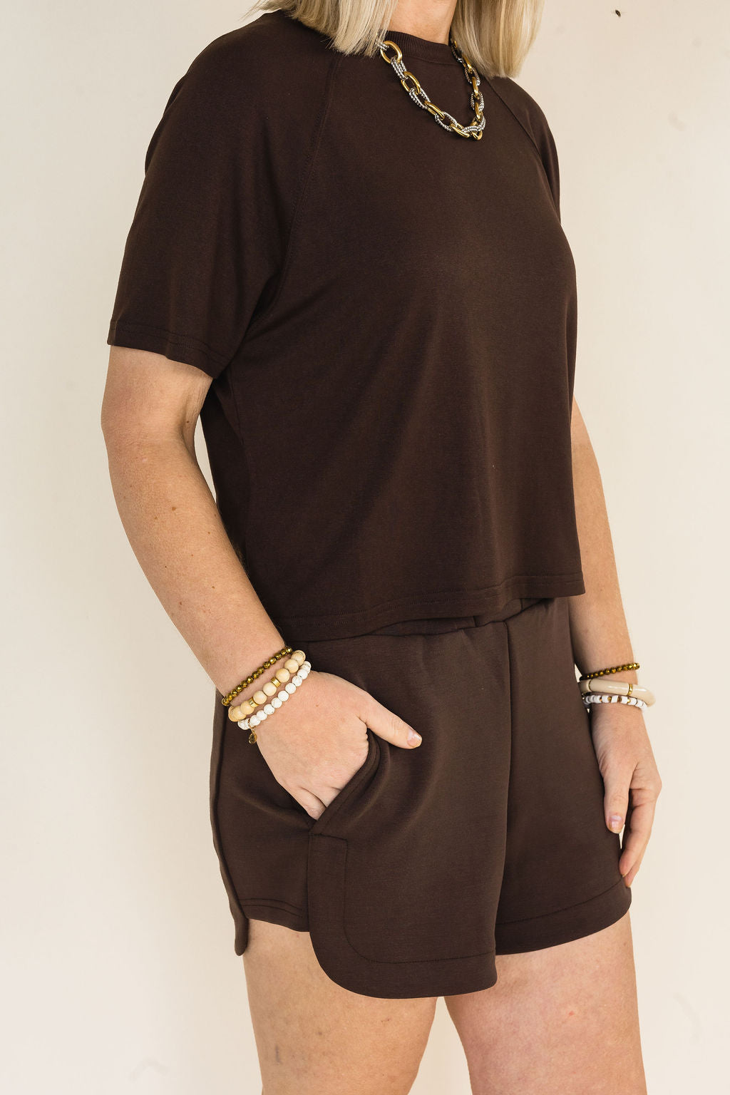 A woman wearing a coffee short-sleeve t-shirt and matching coffee shorts with side pockets, accessorized with gold chain and pearl bracelets, against a plain off-white background.