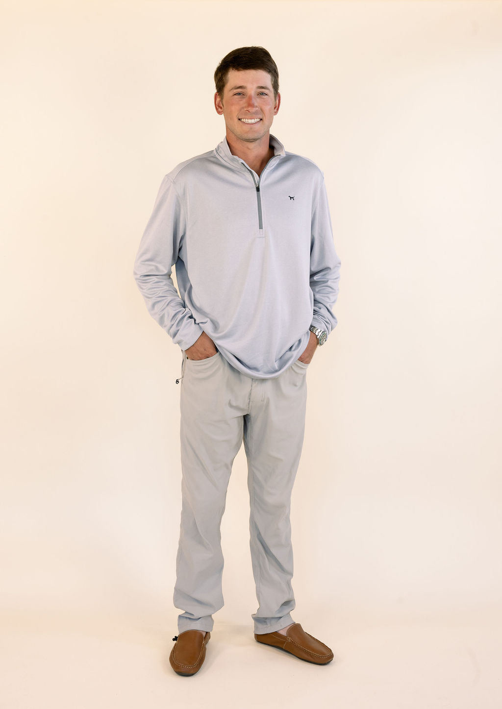 A man wearing a light grey quarter-zip long-sleeve quarter zip and khaki pants, standing against a plain cream-colored studio background.