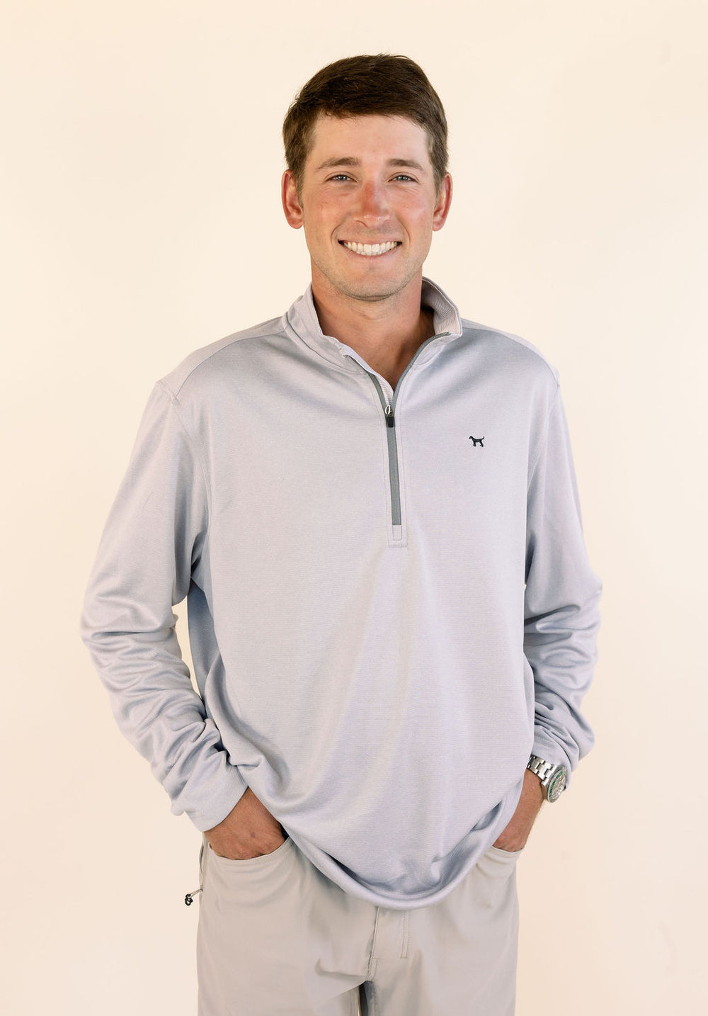 A smiling man wearing a light gray quarter-zip long-sleeve performance pullover and khaki pants, standing against a plain cream background.