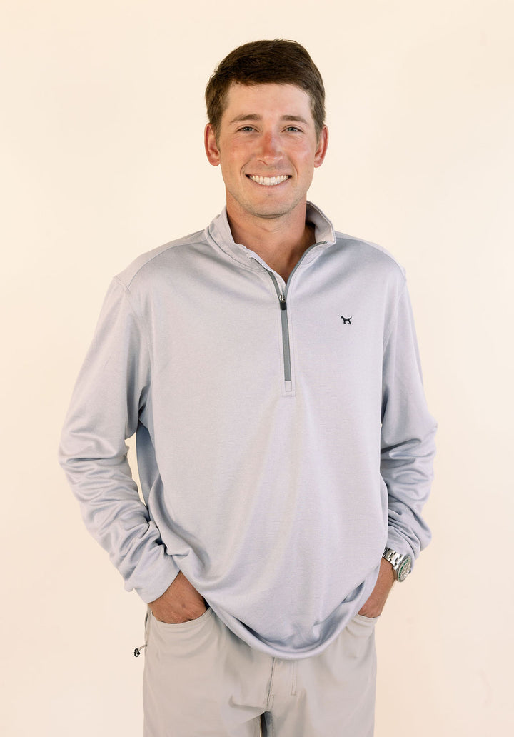 A smiling man wearing a light gray quarter-zip long-sleeve performance pullover and khaki pants, standing against a plain cream background.