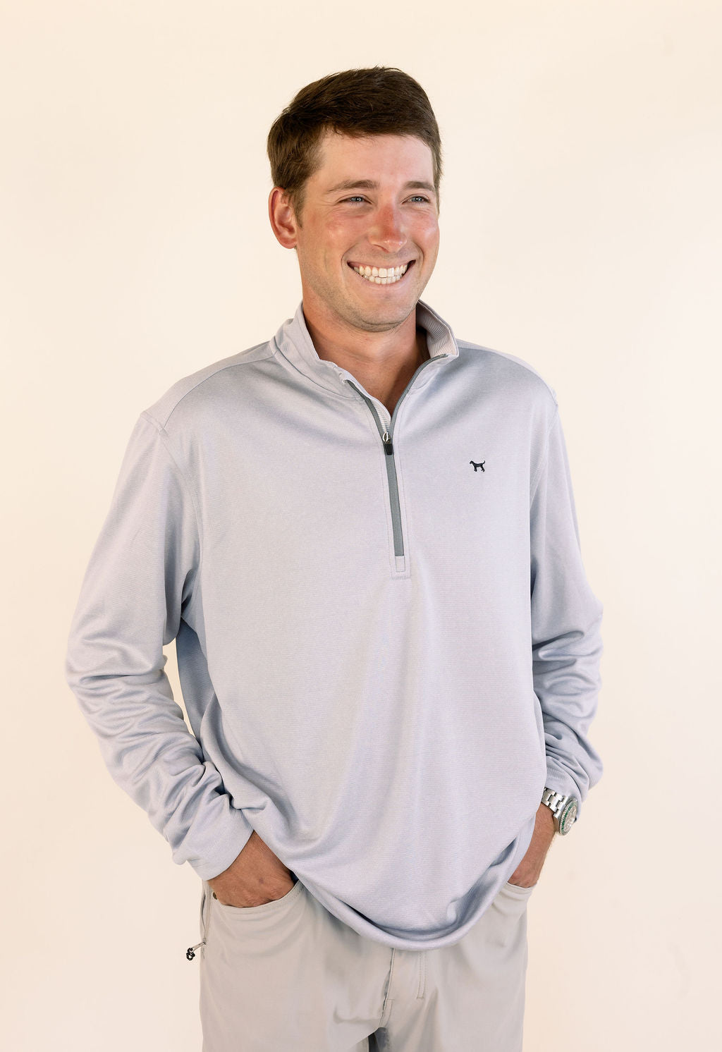 A smiling man wearing a light gray quarter-zip long-sleeve performance pullover and khaki pants, standing against a plain cream background.