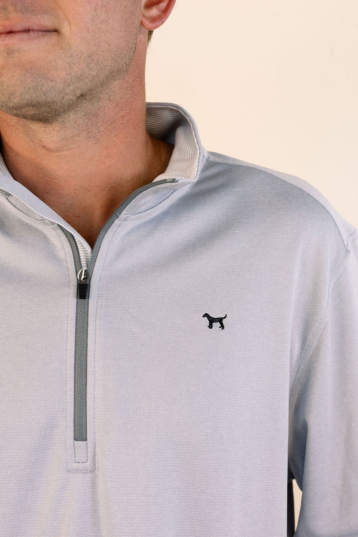 Close-up of a man wearing a light gray quarter-zip pullover with a small black dog logo on the left chest, set against a plain light beige background.