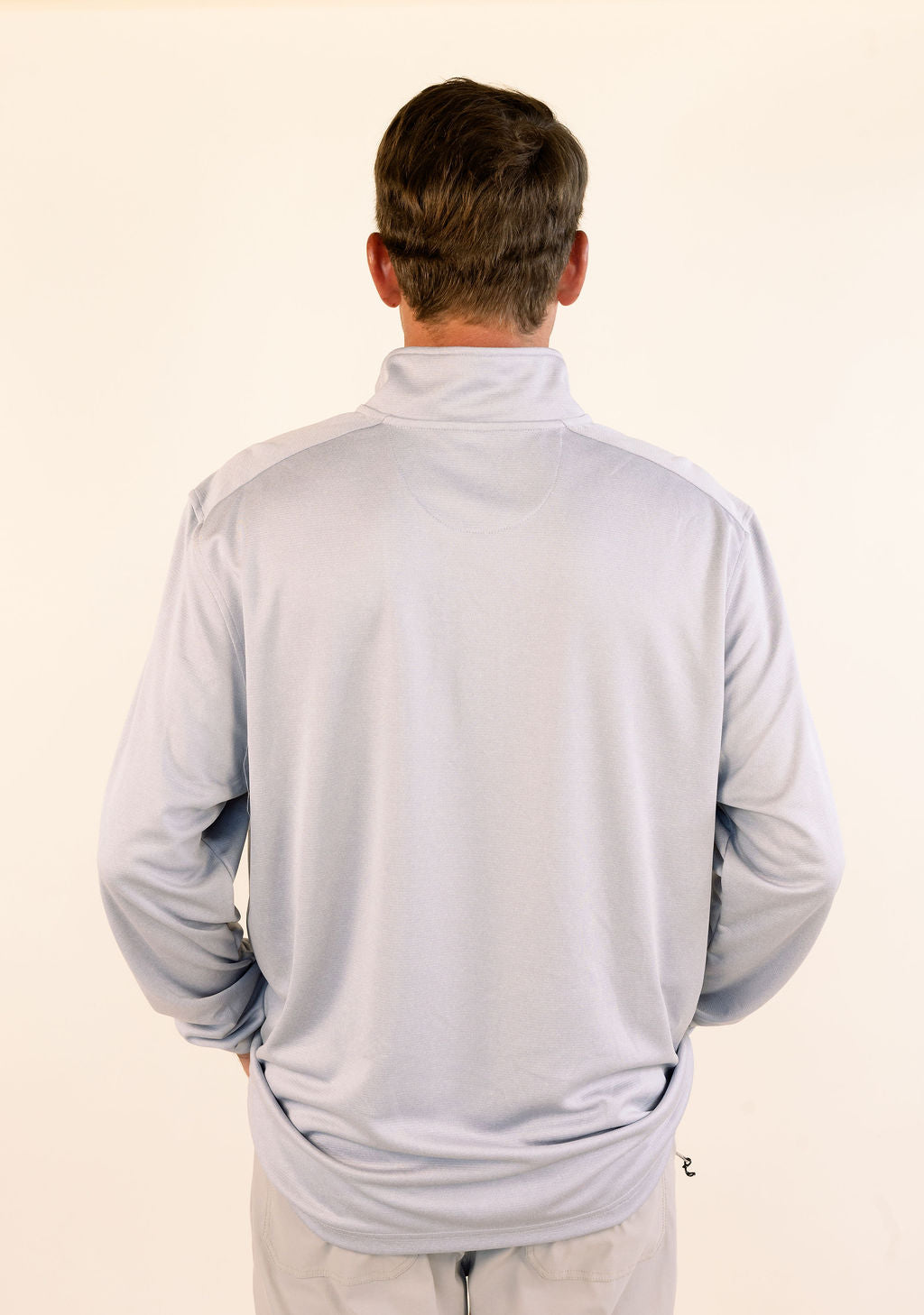 A man viewed from behind wearing a light gray long-sleeve athletic quarter-zip pullover and khaki pants, standing against a plain off-white background.