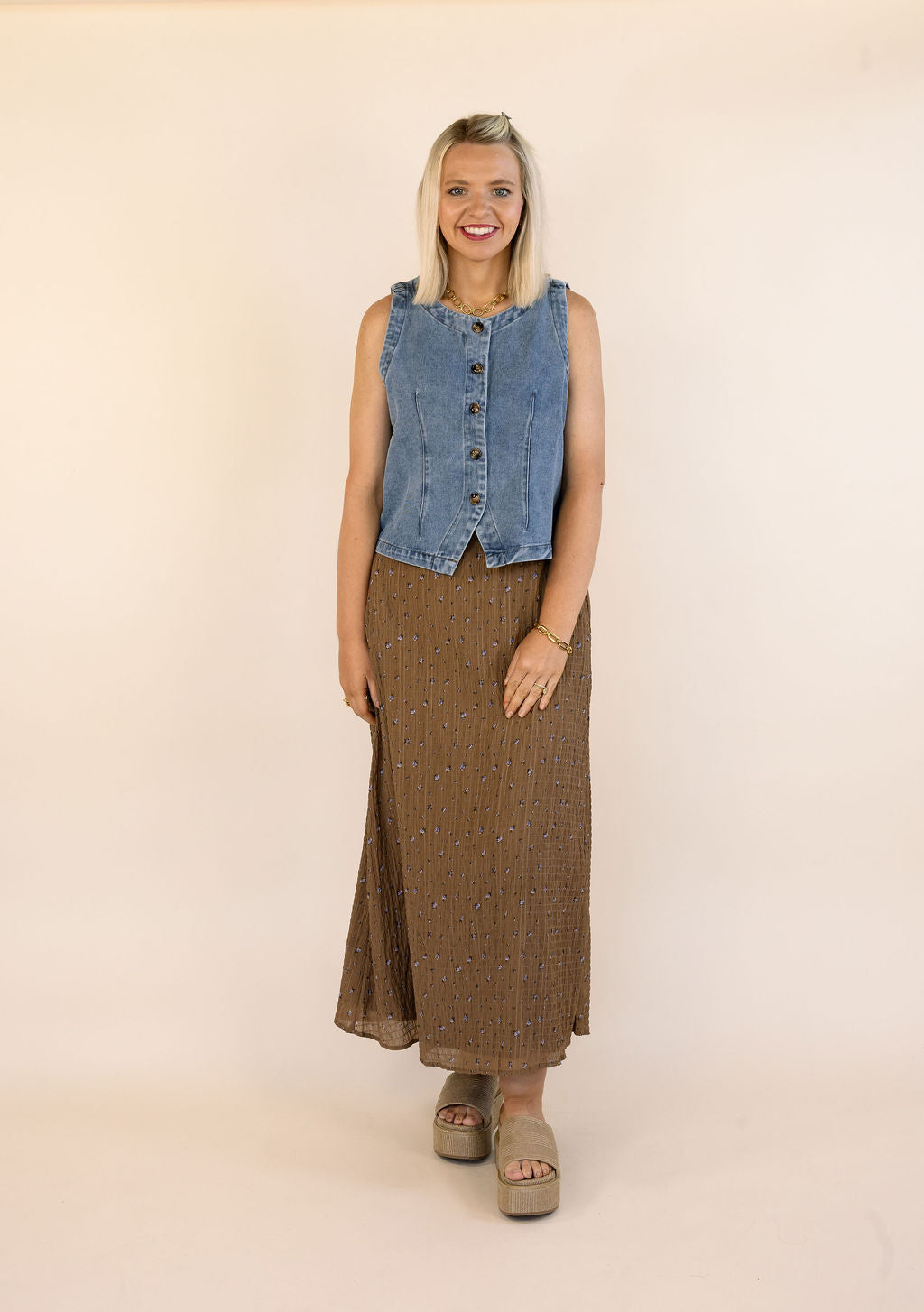 A woman modeling a sleeveless blue denim vest with a button-front design and a brown patterned maxi skirt, standing against a plain beige studio backdrop.