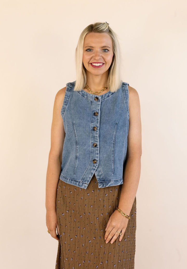 A smiling blonde woman wearing a sleeveless light blue denim vest with dark buttons over a brown patterned skirt, standing against a plain beige background.