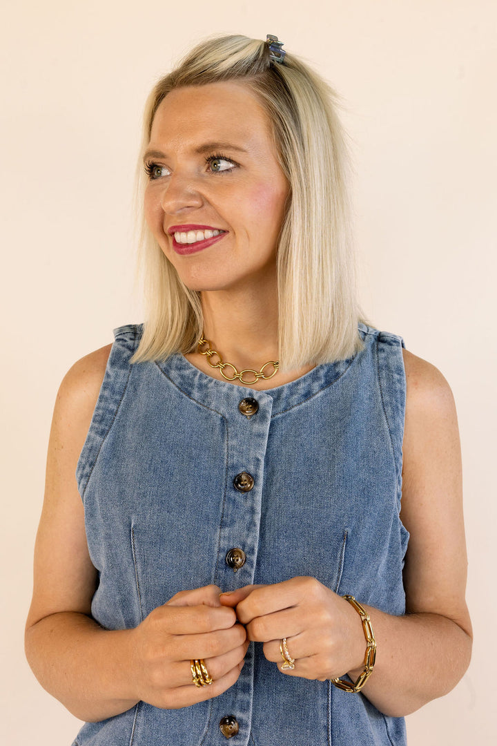 A blonde woman wearing a sleeveless blue denim vest with brown buttons, styled with gold jewelry against a solid beige background.