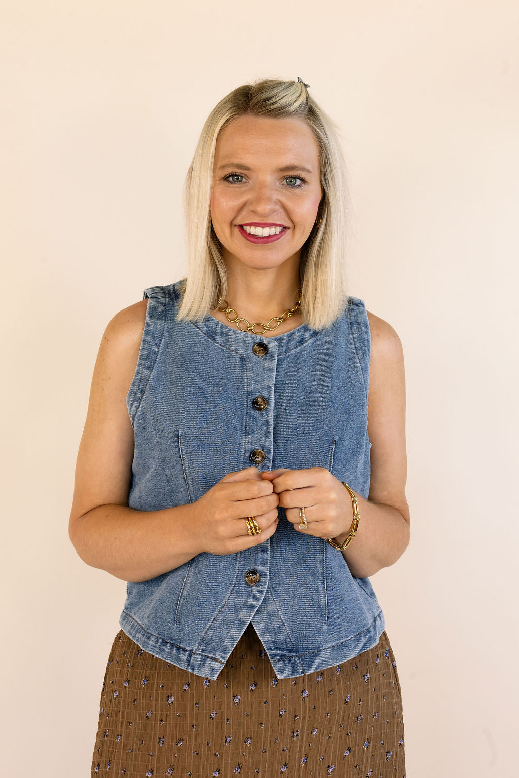 A smiling blonde woman wearing a sleeveless blue denim vest with a button-down front and a brown patterned skirt, standing against a plain beige background.