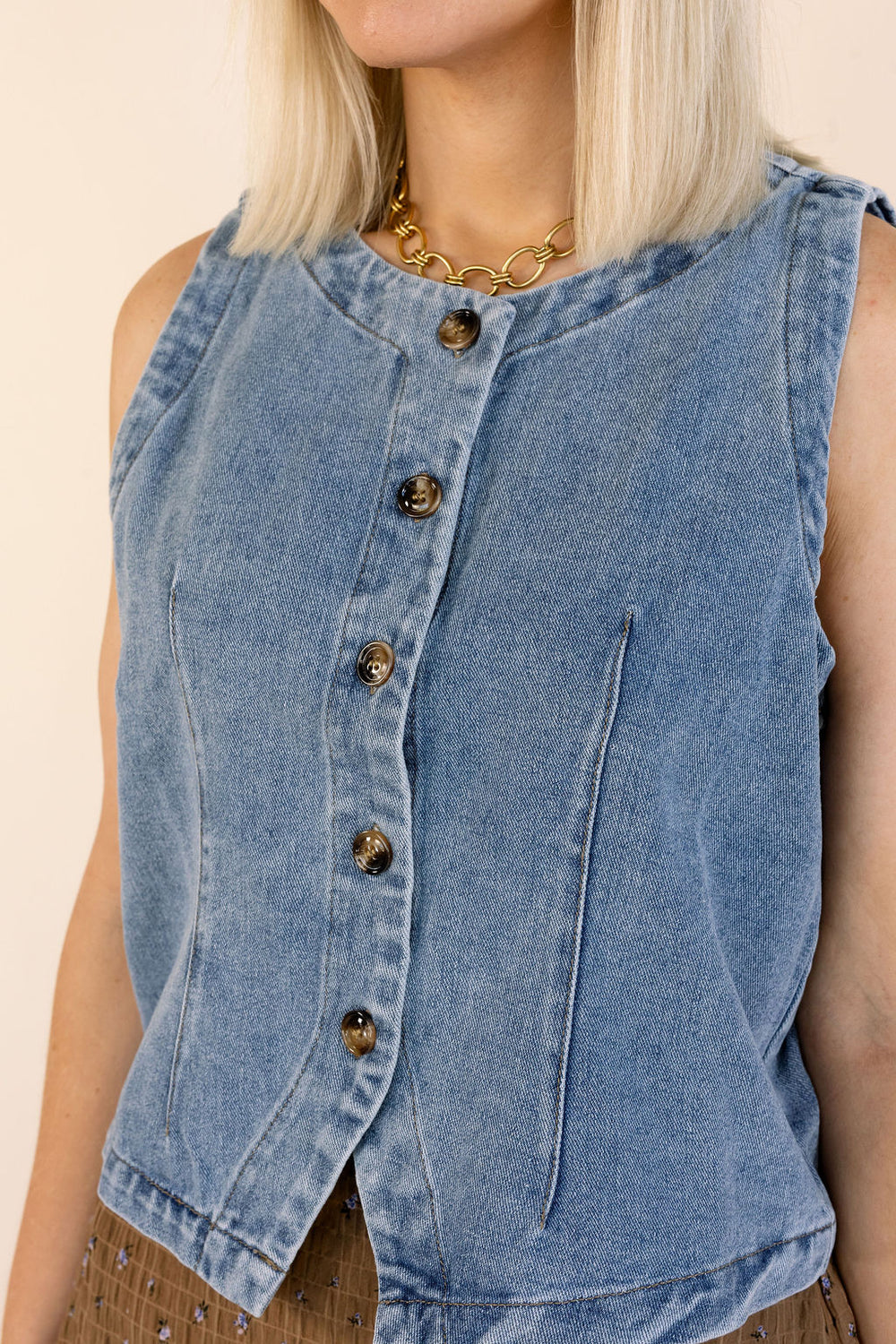 A close-up of a model wearing a sleeveless light blue denim vest with a button-up front and decorative dark buttons.